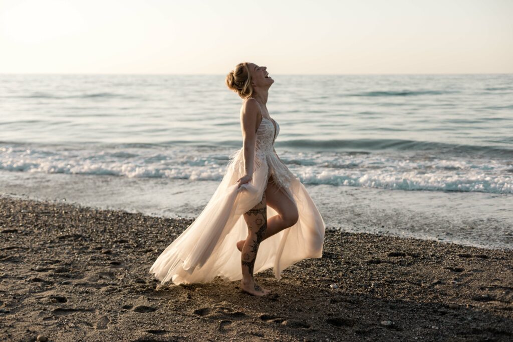 Fröhliche Braut tanzt und baded im Sonnenuntergang Licht goldene Stunde Strand Fotoshooting